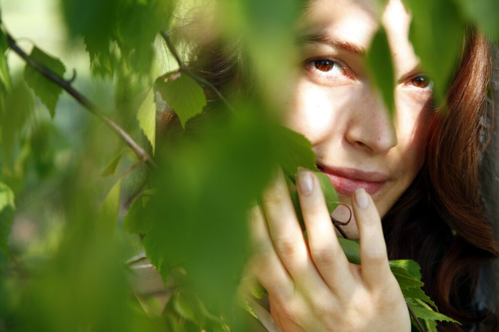 Karolina Kumor-yoniversal care-zioła-olejki-do-pielęgnacji intymnej-olejki do masażu piersi-olejki ziołowe dla kobiet-brzoza-właściwości brzozy-na zdjęciu portret kobiety, która przysłonięta jest listkami i gałązkami brzozy