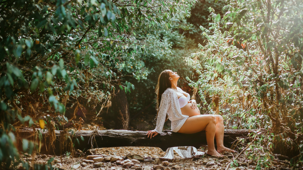 Warsztaty dla kobiet, Yoniversal Care, Karolina Kumor, Narodziny ciąża poród przygotowania, na zdjęciu Kobieta w ostatnim miesiącu ciąży, siedząca w lesie na przewróconym pniu drzewa, trzyma się za brzuch i z zamkniętymi oczami wyciąga głowę do słońca