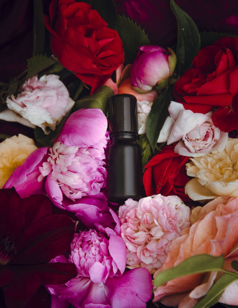 Na zdjęciu mała czarna butelka o pojemnoścl 10 ml. Butelka zawiera perfumy. Leży pośród kolorowych, pięknych kwiatów piwonii i róż.